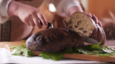 Geleneksel kostümlü bir kadının, yeni pişmiş, dumanı tüten bir somun ekmeği yarıya keserken çekilen resmi.