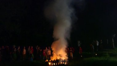 Bir yaz gecesinde yanan ateşin yanında duran bir sürü insanın el kamerasıyla geniş açıdan çekilen görüntüsü. Kamera dumanı takip ediyor.