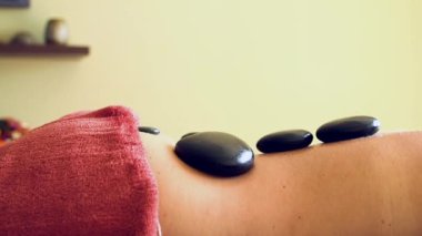Kadın kısmen Havluyla kaplanmış Masaj Masasında Yatıyor. Hot Stones arkada. Sıcak Taş Masajı, masaj terapisinin bir türüdür. Vücut boyunca kasları gevşetmek ve gevşetmek için kullanılır.