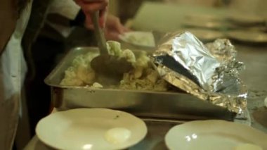 Elinde Slotted Spoon olan bir kadın Sovyet-Dönem kantinindeki bir tabağa hamur tatlısı koyar. 80 'lerin Stili Öğle Yemeği - Yağlı bezle kaplı Masalarda Kesik Cam Bardaklardan İçme.