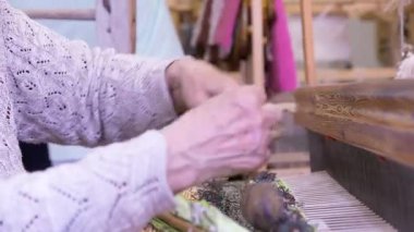 Geniş dokuması olan yaşlı bir kadın renkli, dekoratif bir duvar halısı ya da halısı dokuyor. Dokuma bir tekstil üretim metodudur. Bir kumaş veya kumaş oluşturmak için iki iplik dizisi birbirine karışır..