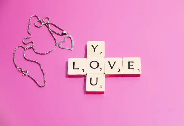 Scrabble harfleri Love You 'yu pembe arka planda bir kalp kolyesiyle heceliyor. Sevgililer Günü.