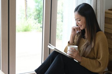 Bir kadın elinde bir defter ve bir fincan kahveyle yerde oturuyor...