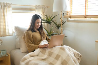 Önünde dizüstü bilgisayarı olan bir kadın yatakta oturuyor. Gülümsüyor ve eğleniyor..