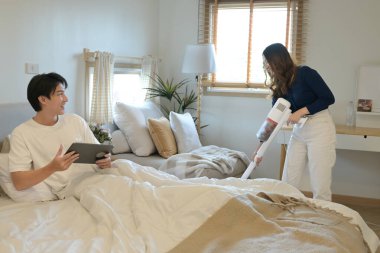 Bir erkek ve bir kadın yatak odasındalar. Adamın elinde tablet var ve kadın süpürge yapıyor..