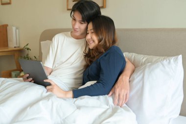 Bir erkek ve kadın, elinde tablet olan bir adamla yatakta uzanıyorlar..