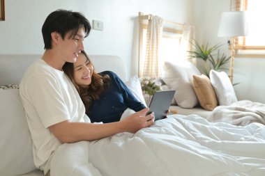 Bir erkek ve kadın, elinde tablet olan bir adamla yatakta uzanıyorlar...