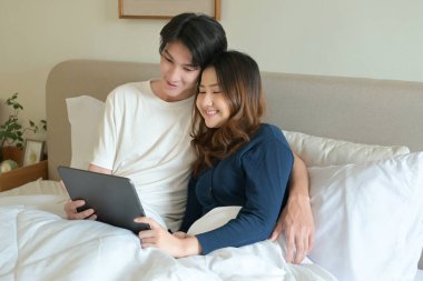 Bir erkek ve kadın, elinde tablet olan bir adamla yatakta uzanıyorlar...