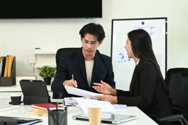 Takım elbiseli iki kişi bir masada arkalarında bir tahtayla konuşuyor. Bir tanesi bir kağıt parçasına yazıyor..