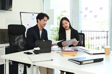 İki kişi bir masada oturuyor, biri bir belge okuyor. Adam takım elbise giyiyor, kadın da takım elbise..