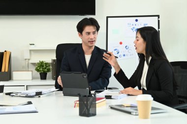 Takım elbiseli iki kişi bir masada arkalarında bir tahtayla konuşuyor. Bir tanesi bir kağıt parçasına yazıyor...