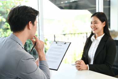 Bir adam ve bir kadın masada oturuyorlar. Adamın elinde bir dosya var ve kadın gülümsüyor. Sahne olumlu ve dostane...