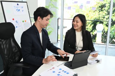 Takım elbiseli iki kişi bir masada arkalarında bir tahtayla konuşuyor. Bir tanesi bir kağıt parçasına yazıyor...