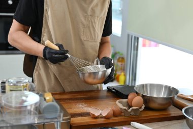Bir adam mutfakta bir pasta yapıyor. Önlük ve eldiven giyiyor. Tezgahta birkaç kase ve kaşık var..