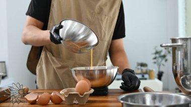 Bir adam kek yapıyor ve bir kaseye sıvı döküyor. Kase ahşap bir masanın üzerinde ve yakınlarda birkaç yumurta ve bir terazi var..