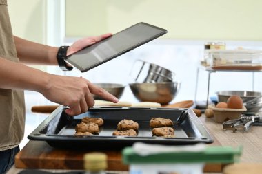 Bir adam elinde tablet tutarken mutfakta yemek pişiriyor. Tableti yemek tarifini bulmak için kullanıyor ya da yemek pişirme kursunu takip ediyor...