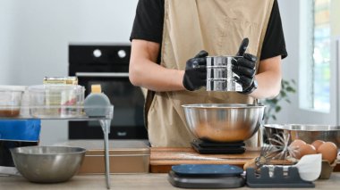 Bir adam kek yapıyor ve unu elemek için elek kullanıyor. Mutfak çeşitli kâseler, kaşıklar ve diğer aletlerle donatılmıştır. Atmosfer sakin ve odaklı...