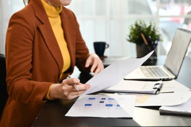 Bir kadın önünde elinde kalem ve kağıtlarla bir masada oturuyor. Kağıtlara yazıyor ve işine odaklanmış durumda...