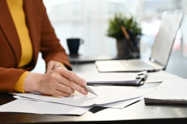 Bir kadın önünde elinde kalem ve kağıtlarla bir masada oturuyor. Kağıtlara yazıyor ve işine odaklanmış durumda...