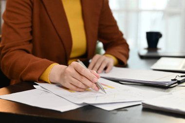 Bir kadın önünde elinde kalem ve kağıtlarla bir masada oturuyor. Kağıtlara yazıyor ve işine odaklanmış durumda..