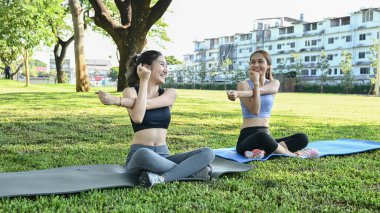 İki kadın parkta yoga yapıyor. Bir tanesi gülümsüyor. Diğer kadın yerde oturuyor..