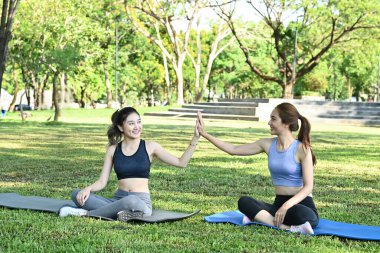 Spor kıyafetli iki kadın parkta bir paspasın üzerinde oturuyor, biri diğerine beşlik çakıyor..
