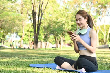 Parktaki bir paspasın üzerinde oturan bir kadın telefonuna bakıyor. Mavi bir atlet ve siyah bir pantolon giyiyor..