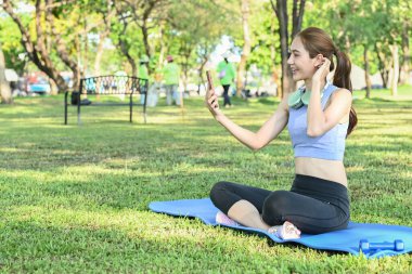 Bir kadın parkta bir paspasın üzerinde oturmuş selfie çekiyor. Mavi bir atlet ve siyah bir pantolon giyiyor..