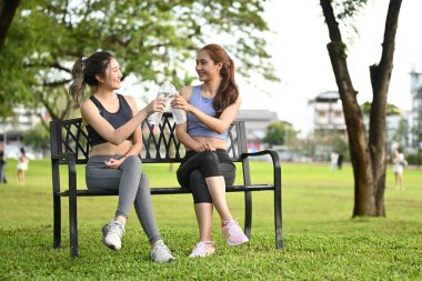 Spor kıyafetleriyle parkta bir bankta oturan iki kadın. Biri elinde bir şişe su tutuyor. Sahne rahat ve sıradan...