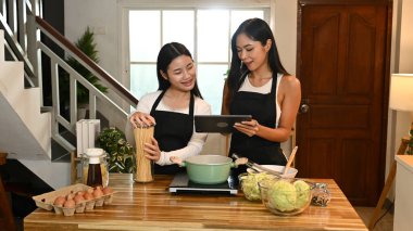 İki kadın mutfakta birlikte yemek pişiriyor. Bir tanesi elinde bir tablet tutarken diğeri elinde bir saksı tutuyor...