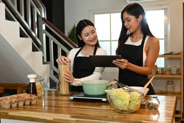 İki kadın mutfakta birlikte yemek pişiriyor. Bir tanesi elinde bir tablet tutarken diğeri elinde bir saksı tutuyor..