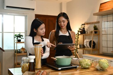 İki kadın mutfakta birlikte yemek pişiriyor. Bir tanesi elinde bir tablet tutarken diğeri elinde bir saksı tutuyor...