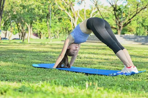 Bir kadın parkta mavi bir mindere yoga pozu veriyor. Gülümsüyor ve eğleniyor...
