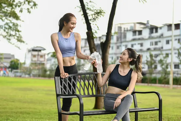Spor kıyafetleriyle parkta bir bankta oturan iki kadın. Biri elinde bir şişe su tutuyor. Sahne rahat ve sıradan...
