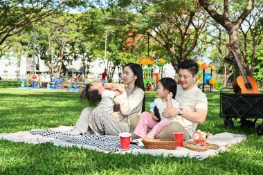 Dört kişilik bir aile parkta bir battaniyenin üzerinde oturmuş pikniğin tadını çıkarıyor..