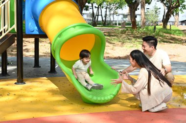 Bir aile kaydırağın önünde duruyor. Ebeveynler izlerken çocuk kaydırağın üzerinde oturuyor. Sahne mutlu ve neşeli...