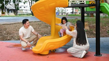 Bir aile kaydırağın önünde duruyor. Ebeveynler izlerken çocuk kaydırağın üzerinde oturuyor. Sahne mutlu ve neşeli...