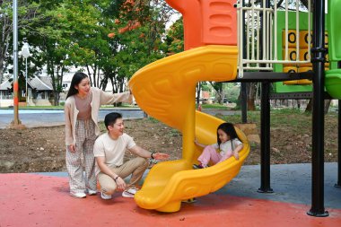 Bir aile kaydırağın önünde duruyor. Ebeveynler izlerken çocuk kaydırağın üzerinde oturuyor. Sahne mutlu ve neşeli...