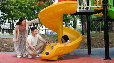 Bir aile kaydırağın önünde duruyor. Ebeveynler izlerken çocuk kaydırağın üzerinde oturuyor. Sahne mutlu ve neşeli...