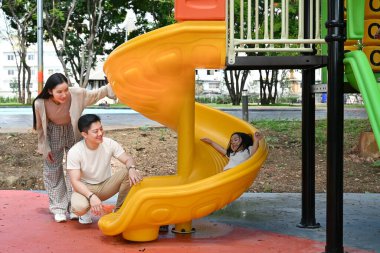 Bir aile kaydırağın önünde duruyor. Ebeveynler izlerken çocuk kaydırağın üzerinde oturuyor. Sahne mutlu ve neşeli...