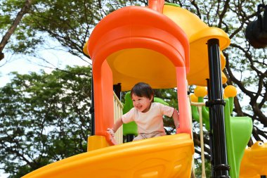 Küçük bir çocuk sarı ve turuncu bir oyun yapısında oynuyor. Çocuk gülümsüyor ve eğleniyor..