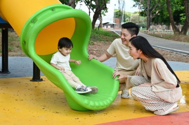 Bir aile kaydırağın önünde duruyor. Ebeveynler izlerken çocuk kaydırağın üzerinde oturuyor. Sahne mutlu ve neşeli..