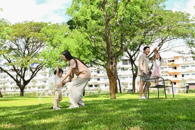 Başka bir baba izlerken bir anne çocuğunu kucağında tutuyor. Çocuklar parkta oyun oynuyorlar. Sahne mutlu ve neşeli...