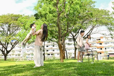 Başka bir baba izlerken bir anne çocuğunu kucağında tutuyor. Çocuklar parkta oyun oynuyorlar. Sahne mutlu ve neşeli..