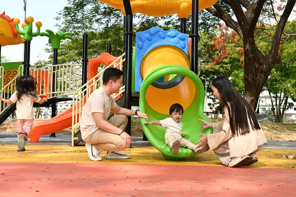 Bir aile kaydırağın önünde duruyor. Ebeveynler izlerken çocuk kaydırağın üzerinde oturuyor. Sahne mutlu ve neşeli...