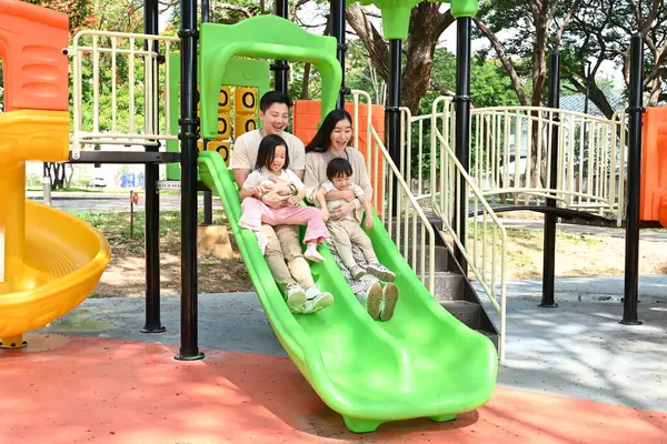 Dört kişilik bir aile parkta yeşil bir slaytta eğleniyor. Sahne mutlu ve eğlenceli, çünkü aile birlikte kaliteli zaman geçiriyor ve eğleniyor..