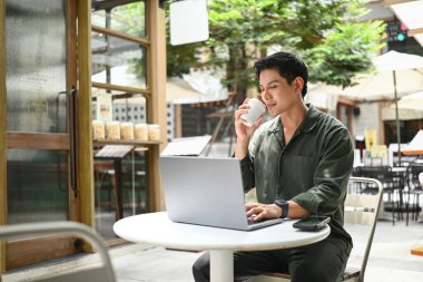 Bir adam dışarıda küçük bir masada oturuyor, dizüstü bilgisayarına dalmış, aynı zamanda bir fincan kahvenin tadını çıkarıyor..