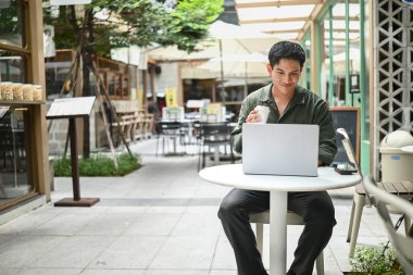 Bir adam dışarıda küçük bir masada oturuyor, dizüstü bilgisayarına dalmış, aynı zamanda bir fincan kahvenin tadını çıkarıyor..