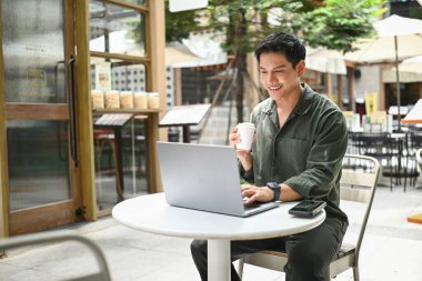 Bir adam dışarıda küçük bir masada oturuyor, dizüstü bilgisayarına dalmış, aynı zamanda bir fincan kahvenin tadını çıkarıyor..
