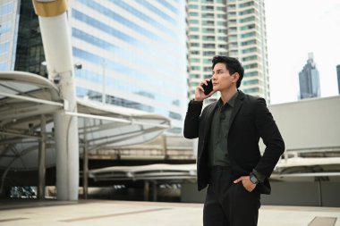 Takım elbiseli bir adam dışarıda dururken cep telefonuyla konuşuyor. Keyfi yerinde ve profesyonel olarak giyinmiş...